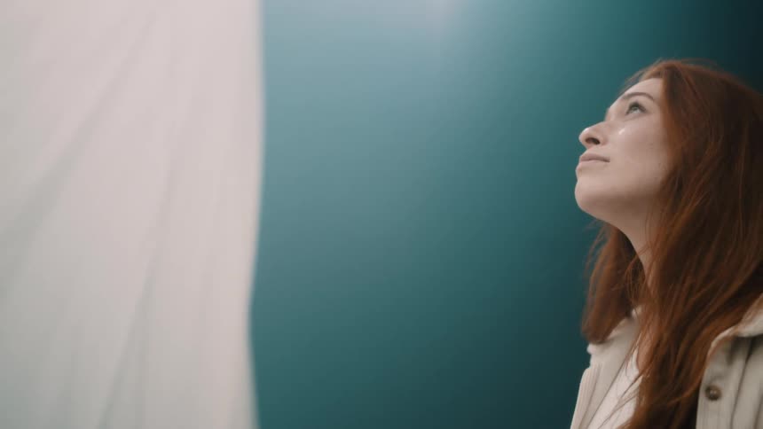 A young woman stands inside a bright room her long hair falling freely as she gazes upward toward the ceiling