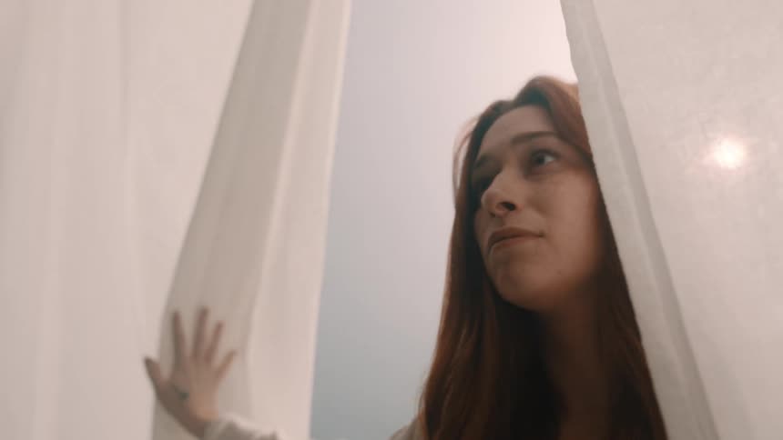 A red haired woman with a small cross tattoo on her hand pulls back sheer white curtains looking up as she walks forward