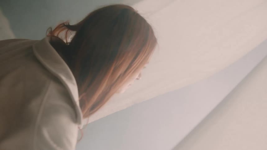 A woman with long auburn hair stands by a window gently parting sheer white curtains to look outside warm hazy sunlight streams into the room creating a soft ethereal glow as the camera tilts upward