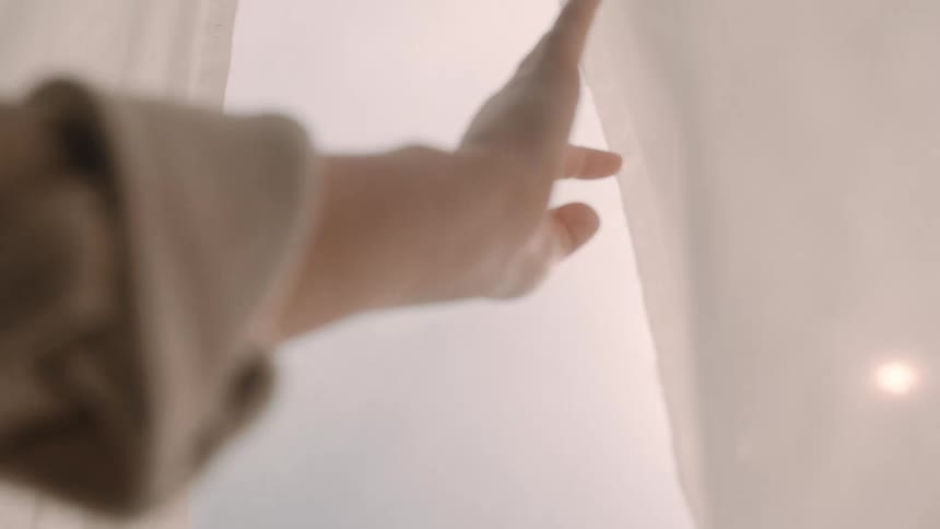 A hand gently draws aside a sheer white curtain letting in soft diffused daylight