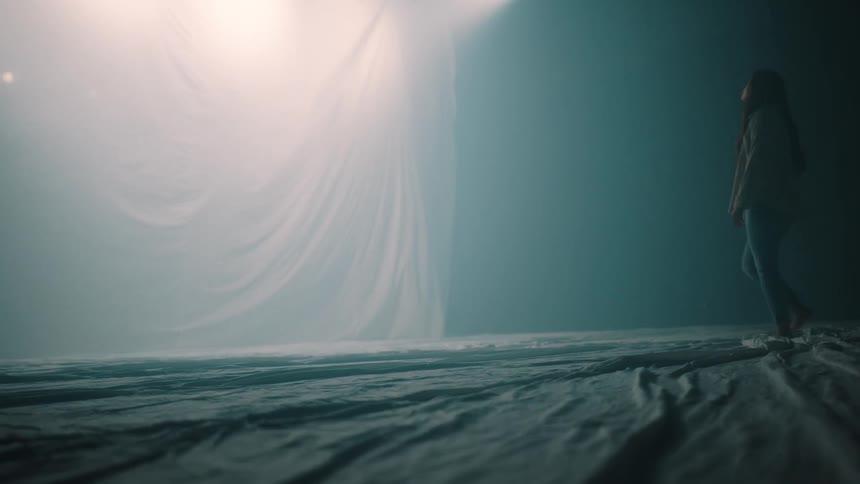 A young woman walks through a large tent filled with white smoke moving toward another section separated by a white linen panel bright light reflecting softly off the fabric as she approaches