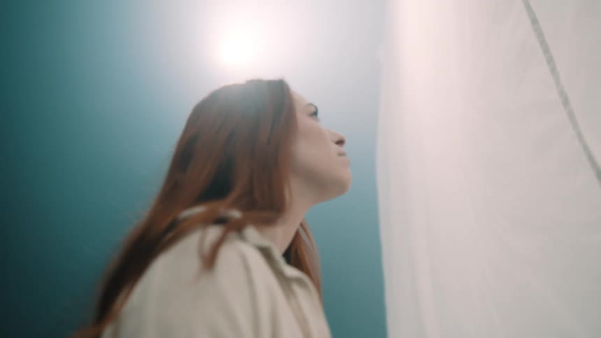A view from below shows a young woman with long red hair wearing a tan shirt and jeans as she looks upward toward a bright light