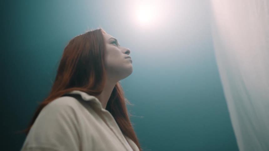 Woman standing under a light at the top of a blue wall looks up at hanging white fabric in front of her