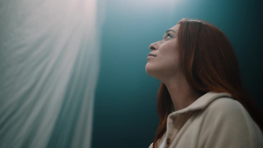 A red haired woman looks up with a serene expression as a white curtain blows from a gentle breeze
