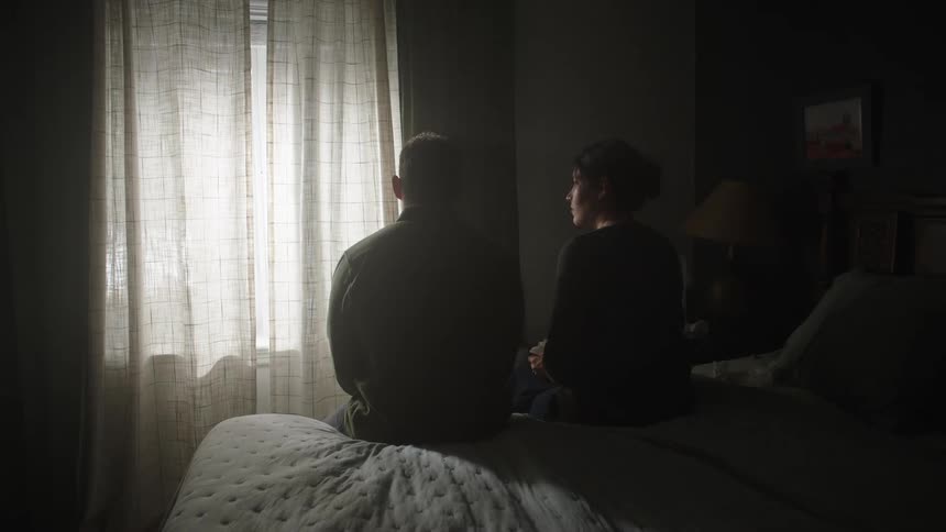 Couple sits on the bed watching light pass through translucent curtains near the window