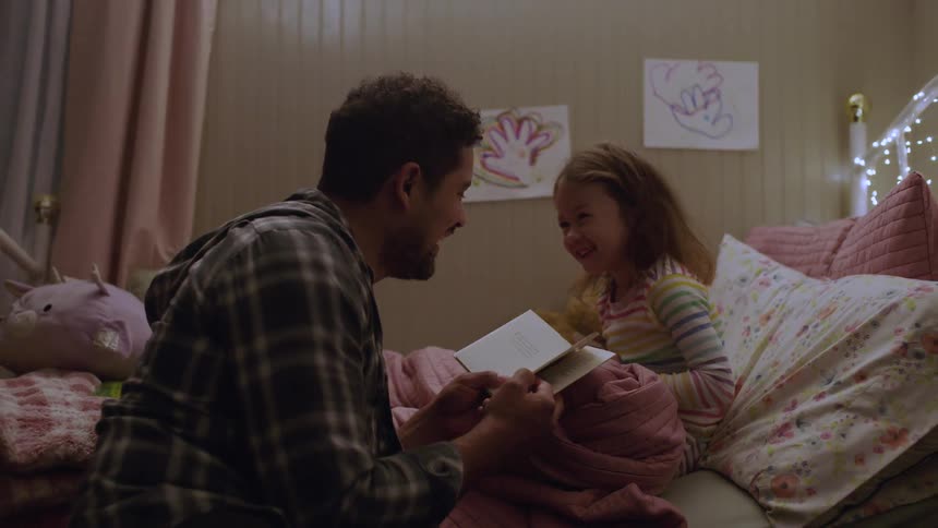 Little girl sits in bed laughing as her father reads a book