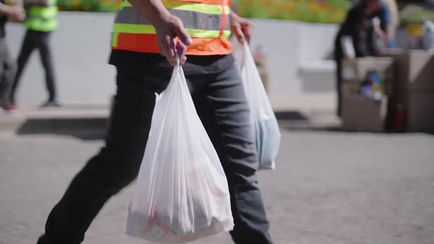 A close up shot shows a volunteer in a bright orange safety vest carrying two white plastic bags filled with groceries and placing them into the trunk of a car at a food distribution site