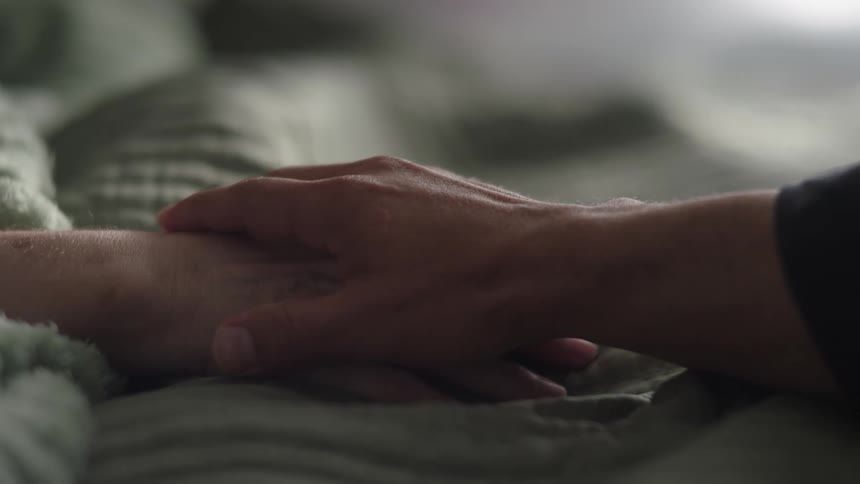 Young adult man holding the hand of an elderly woman lying down on a bed indoors during day time