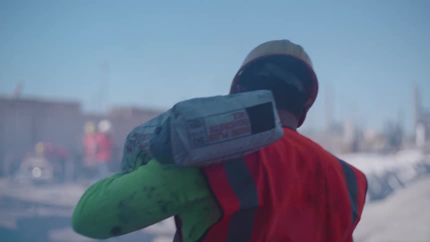 A construction worker in a highvisibility orange vest and hard hat walks through a bright sunlit site while carrying a heavy bag on their shoulderthe scene is captured from a following perspective with a shallow depth of field blurring the industri