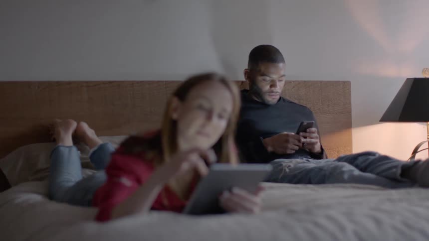 Two people are on the bed using devices