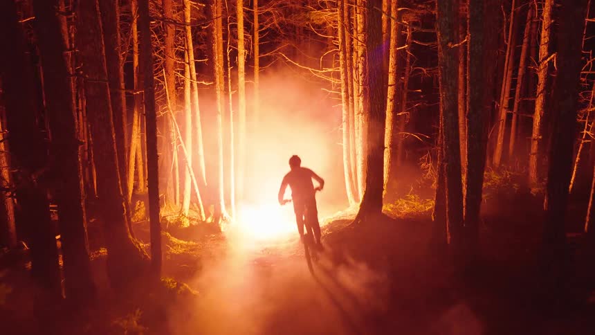 A mountain biker rides away from the camera through a dark forest at night illuminated by a bright orange light source and misty fog that creates long shadows among the tall pine trees as the person disappears into the distance