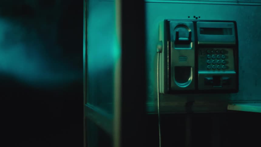A closeup shot shows an old public payphone mounted inside a glass booth at night illuminated by a moody teal light