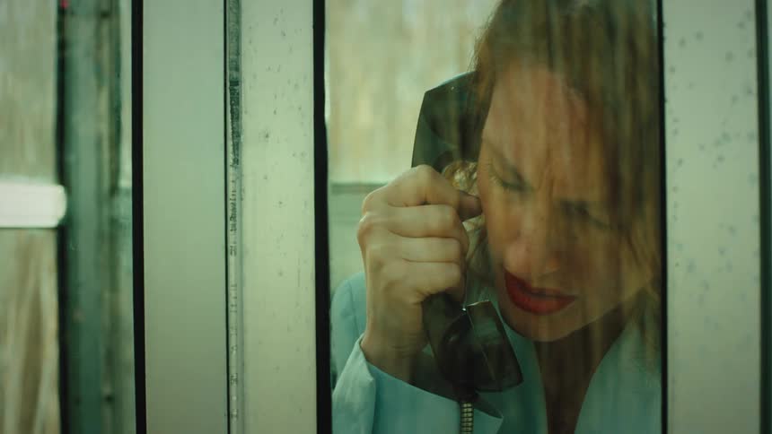A distressed woman speaks on a telephone inside a glass booth