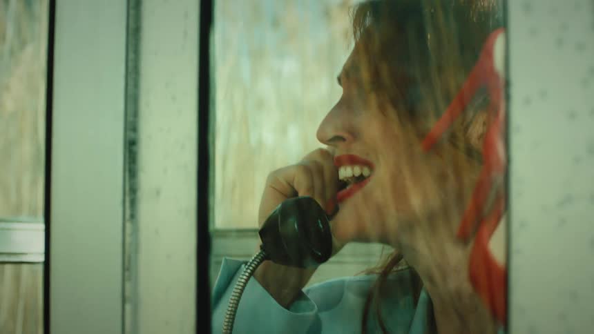 A woman with red lipstick and a blue jacket shouts into a black handset inside a glass phone booth while holding a pair of red high heels against the window