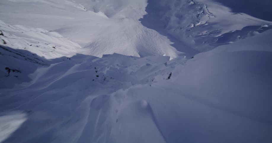 An aerial camera glides over a vast mountain range covered in thick white snow revealing jagged rocky peaks and deep shadows under a clear blue sky during a bright winter day