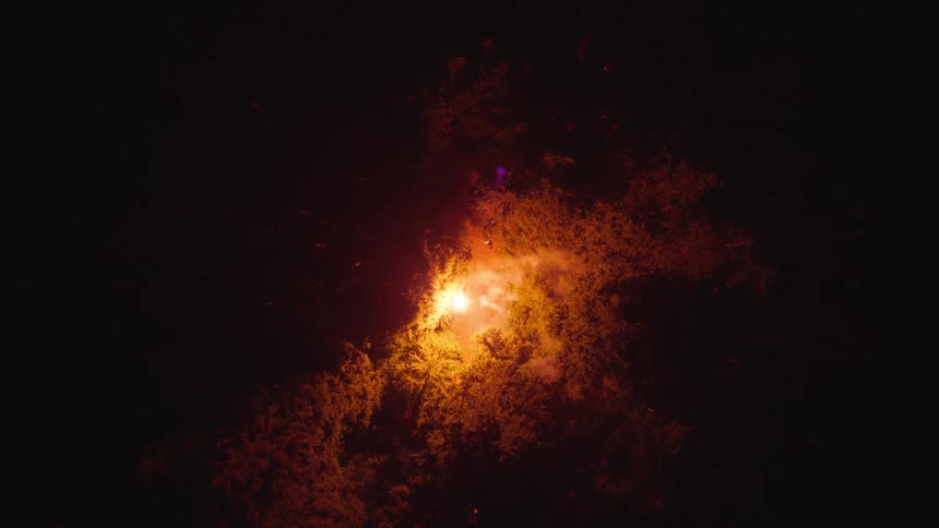 An aerial view ascends over a dark forest illuminated by a bright smoking flare