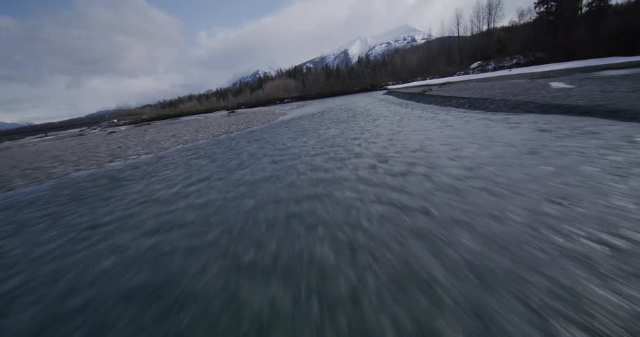 A river runs between rows of winterbare trees its surface cold and still beneath the midday light with a mountain range rising in the background its snowy peaks clear against the bright sky