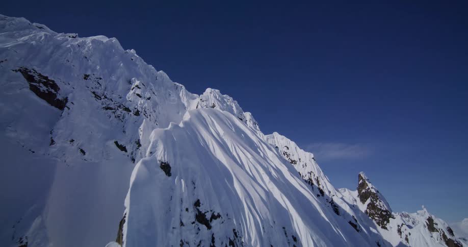 An aerial view reveals a jagged snowcovered mountain ridge with deep vertical fluting and windsculpted peaks under a clear blue sky