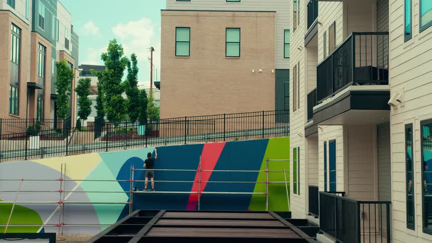 Person painting a geometric mural on an exterior wall using scaffolding between residential buildings