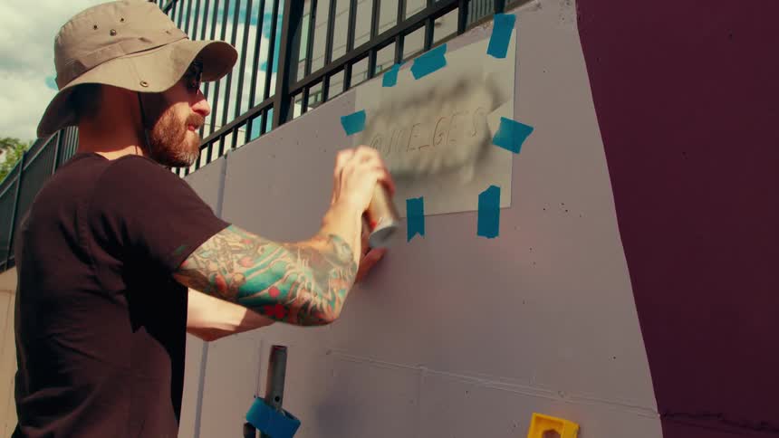 A tattooed man wearing a boonie hat uses a spray can to apply black paint over a paper stencil taped to a concrete wall