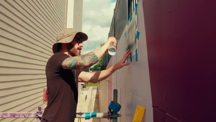 A man with arm tattoos wearing a sun hat and sunglasses uses a spray can to paint over a stencil on a large outdoor wall while standing on scaffolding next to a residential building