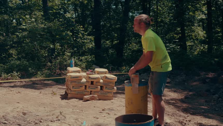 Two men in yellow shirts assembling a cylindrical shape on the ground with a measuring tape and bags in the background