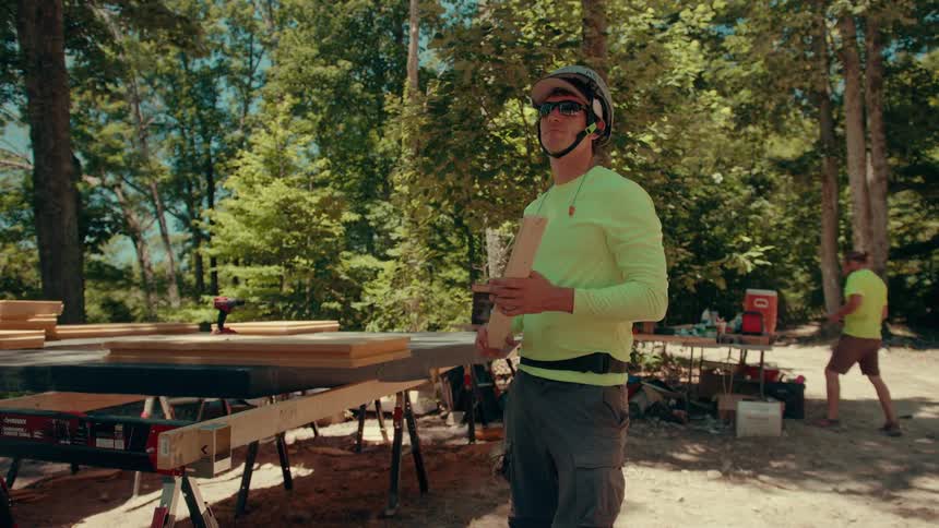 A worker in a highvisibility shirt and safety helmet stands at an outdoor construction site while holding a piece of lumber