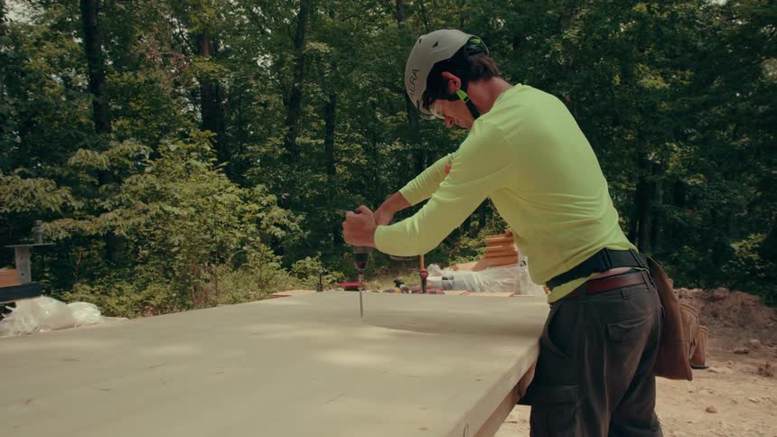 Person wearing helmet and neon shirt using drill on wooden panel in wooded area