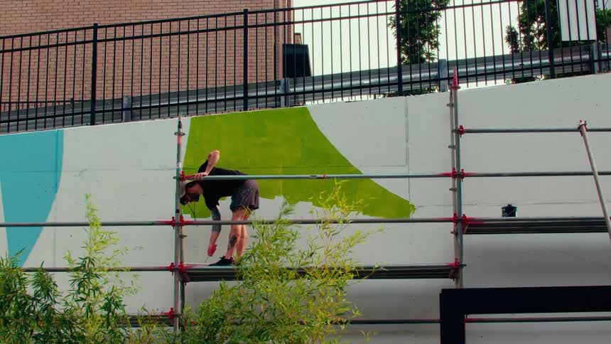 A man wearing a tan sun hat stands on metal scaffolding while painting a bright limegreen section of a large outdoor mural looking forward with focused intent