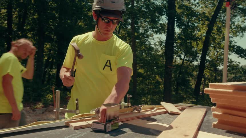 Person in yellow shirt and helmet hammering wooden plank in wooded area with tools around