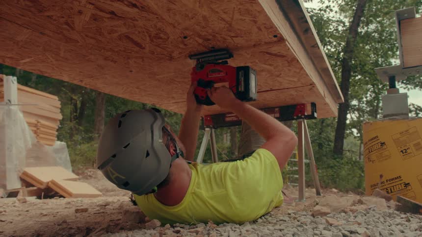 A man wearing a grey helmet and limegreen shirt lies on his back to operate a red power tool against a wooden board looking forward with concentration