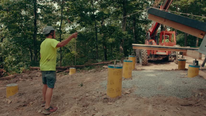 A man in a hardhat moves and guides a cabled counter top on a construction site in a wooded area