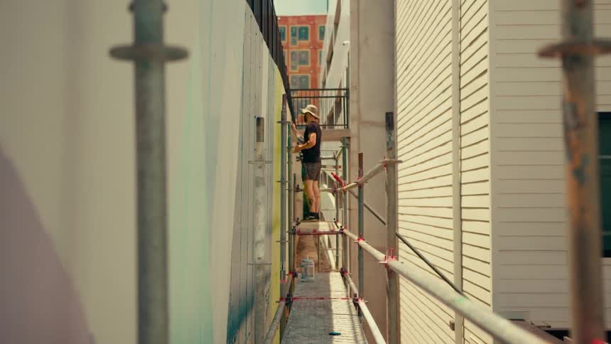 A man wearing a straw hat walks across a narrow metal scaffolding and begins painting a colorful mural on a large outdoor wall located between two buildings during the day