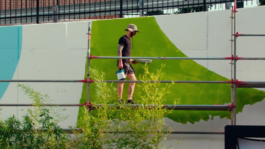 A man in a tan hat walks carrying a paint tray and paint can along a metal scaffold in front of a limegreen section of an outdoor mural