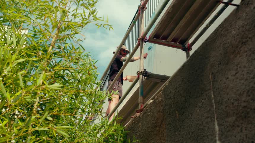 A man wearing a hat and shorts stands on a metal scaffold against a tall concrete wall painting it while green foliage partially frames the left side under a bright cloudy sky