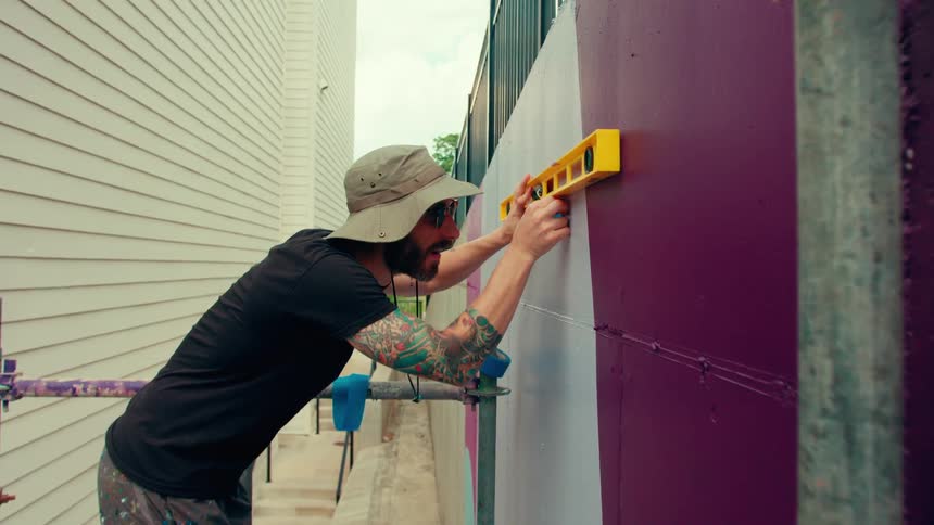 A young man wearing a sun hat stands on a scaffold at midday using a bubble level to mark the wall with steady careful lines