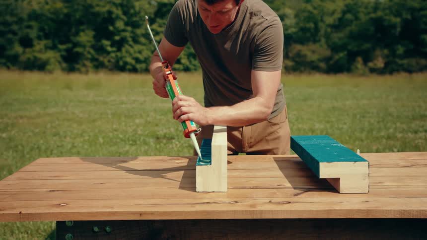 A closeup shot shows a man in a brown tshirt meticulously applying a line of green adhesive from a caulking gun onto a piece of blue construction material outdoors