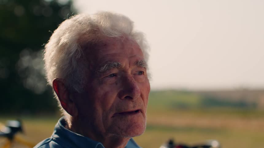 Whitehaired man wearing blue shirt in rural setting with yellow bicycle in the background