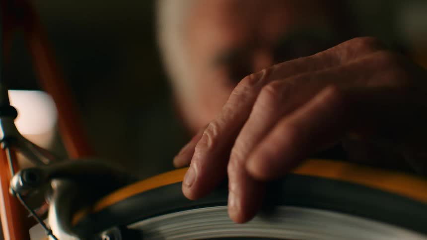 An elderly man works on his bicycle wheel inside his garage leaning over the frame as he makes careful adjustments in the quiet cluttered space