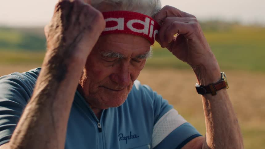 A senior male cyclist stands outdoors in a rural landscape where he unbuckles and removes his yellow helmet before adjusting a red headband on his forehead while looking thoughtfully into the distance during a bright day