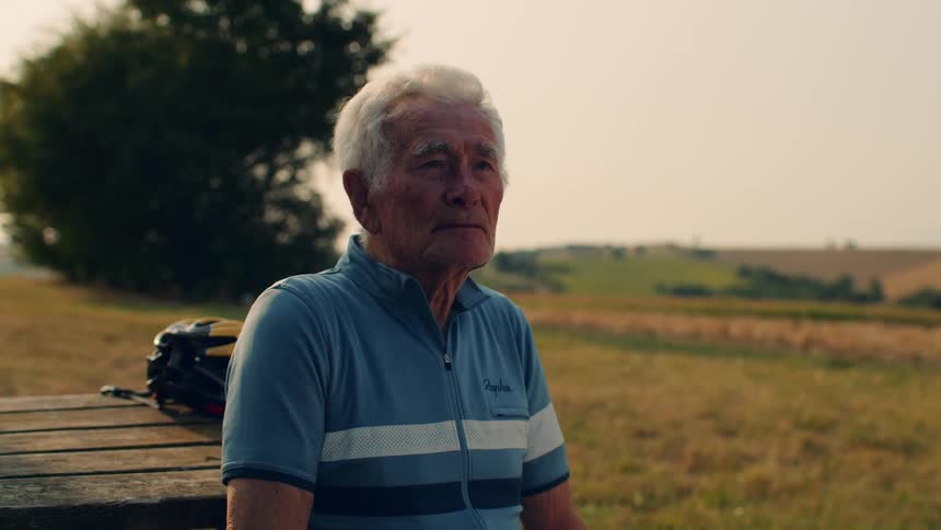 An elderly man in cycling gear sits down on a wooden bench in a field to rest during a sunset