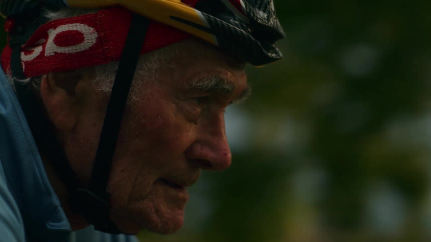 A close up shot shows the profile of an elderly man wearing a cycling helmet and gear as he focuses while riding