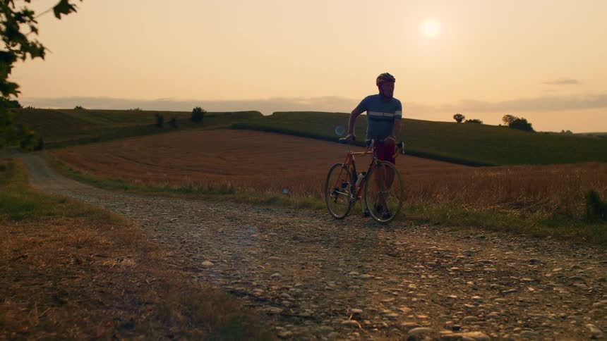 A male cyclist in athletic gear walks his vintage road bike along a rocky gravel path through rolling hills at sunset