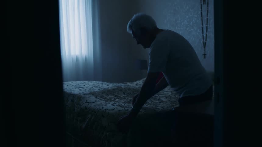 In a somber bluelit bedroom an elderly man gets up from the edge of the bed walks around to smooth the covers and then prepares for his day in solitude