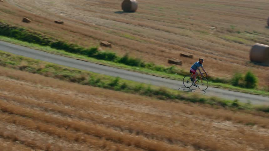 An aerial shot tracks a cyclist as they pedal along a winding country road sweeping past vast sunlit fields dotted with hay bales and sunflowers