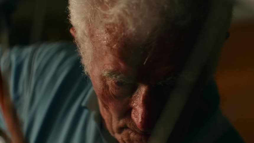 Whitehaired elderly person with focused expression and blue shirt in warmly lit setting