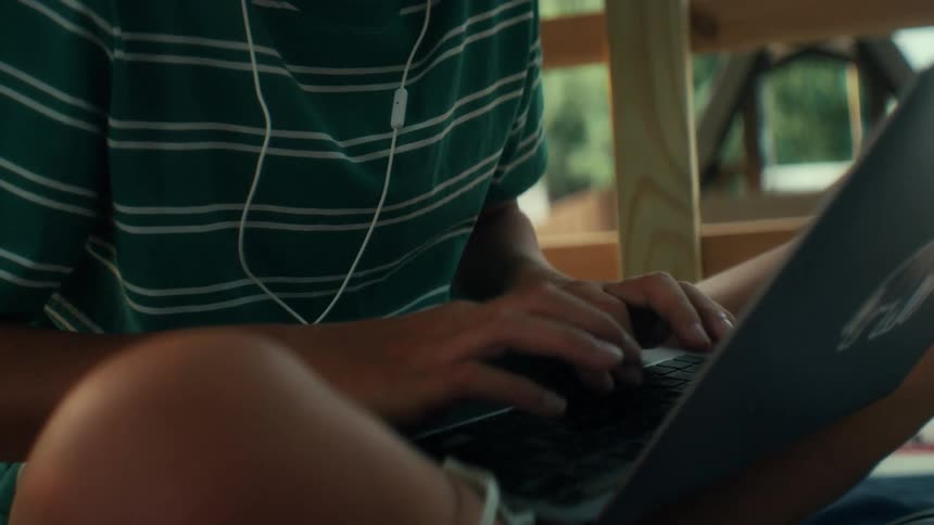 A boy in green and white striped shirt and white earphones typing on laptop doing homework inside naturally lit tree fort
