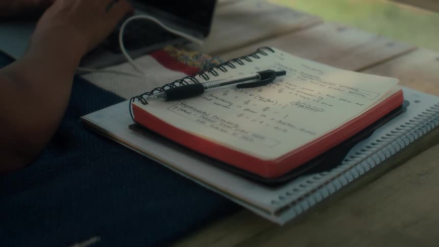 A pen rests on a notebook filled with homework and lecture notes outside in a treefort