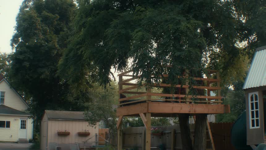 Boy relaxing in a treefort in a residential backyard with houses and fences around it in the neighborhood