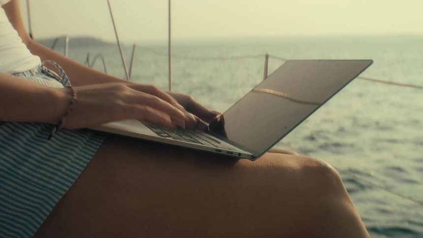 A person seen from the waist down is sitting using a laptop on their lap on the water